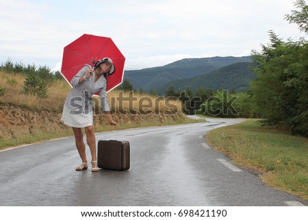 A scene with a beautiful young woman who is posing with a red umbrella and an old - fashioned suitcase on a wet road