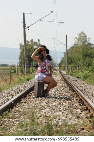 A beautiful young woman is posing with an old - fashioned suitcase on the rails