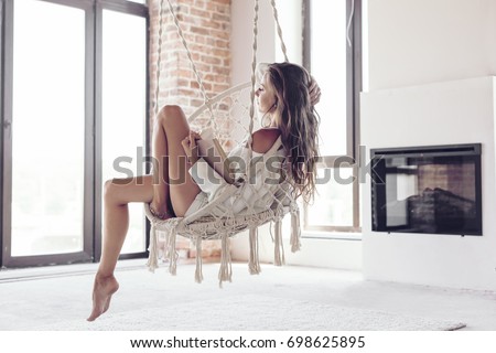 Young woman chilling at home in comfortable hanging chair near fireplace. Girl relaxing and reading book in swing in loft living room with brick walls. Royalty-Free Stock Photo #698625895