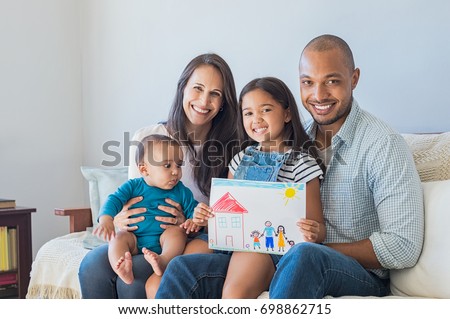 Daughter showing drawing of a happy family in new house. Cute infant looking at colorful drawing of his sister. Happy proud multiethnic parents sitting with children on sofa  and looking at camera. Royalty-Free Stock Photo #698862715