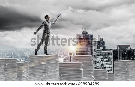 Man in casual wear keeping hand with book up while standing on pile of documents with cityscape and sunlight on background. Mixed media.