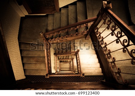 Old and dark squared spiral multi-flight stairway with brown wood and metal handrails seen from the top of the staircase.