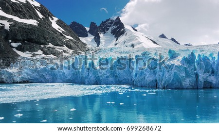 Blue and turquoise colored ice of glacier face with water reflections. Jagged mountains in background of Drygalski Fjord, South Georgia Island in the South Atlantic Ocean.  Royalty-Free Stock Photo #699268672