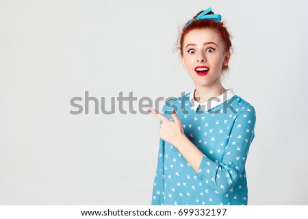 Excited young redhead girl with hair knot pointing her index finger sideways, raising eyebrows and keeping mouth wide opened, showing something surprising on grey copy space wall. Indoor, studio shot