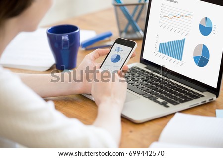 Businesswoman holding smartphone while working with laptop at home office desk, using cross platform responsive design apps for project analysis, analyzing cloud data statistics, close up rear view Royalty-Free Stock Photo #699442570