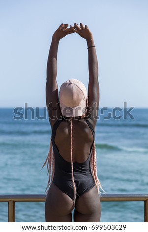 African young girl in a black bathing suit and cap with long pink pigtails posing in the background of the beach and ocean