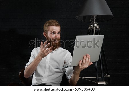 Picture of excited young red-haired male photographer with fuzzy beard dressed in white shirt holding open laptop in front of him and gesturing while making video call using online services