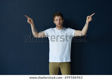 Advertising and promotion concept. Indoor shot of emotional young unshaven hipster dressed in white t-shirt and jeans raising his tattooed arms and pointing fingers in the opposite direction