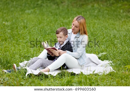 Young mother helping her son child with homework, lying on grass and reading book at park