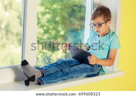 Little boy sits on the windowsill and listening to music through headphones and looking out into the street. The child uses the tablet to watch cartoon, play games, engage in Internet surfing.