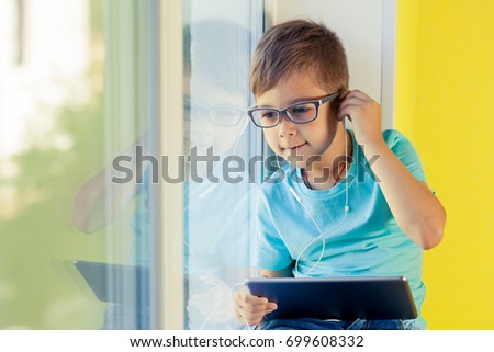 Little boy sits on the windowsill and enjoy listening to music on radio through headphones. Best playlists and new singles. The child uses the tablet to watch cartoon, play games, Internet surfing.