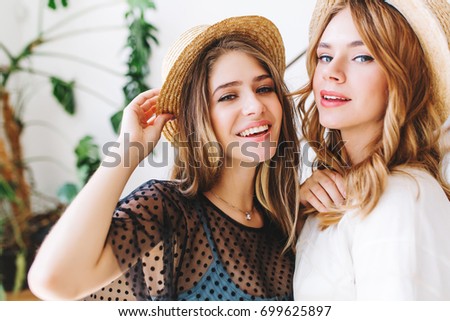 Close-up portrait of wonderful girls with curly hair posing in hats in front of wall with plant. Two young ladies with lovely face expression spending time together and smiling to camera.