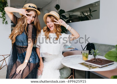 Excited girls in similar straw hats posing with peace sign laughing and fooling around. Indoor portrait of two ladies in good mood dancing in cafe after shopping together. Royalty-Free Stock Photo #699625915