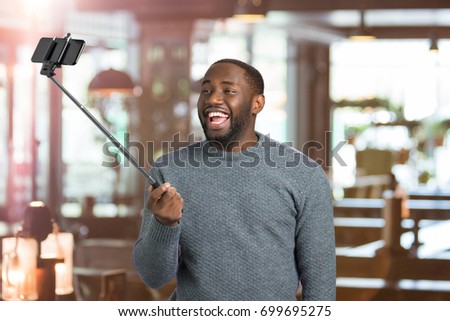 Joyful guy taking picture with selfie stick. Happy young man taking selfie holding monopod on blurred background. Taking sweet moments with selfie stick.