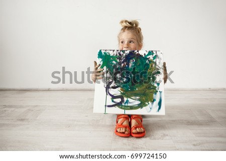 Isolated headshot of Caucasian blonde preschool girl showing picture that she painted. Adorable child holding canvas. Happy childhood concept.