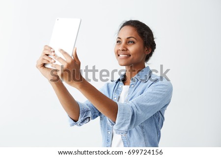 Pretty and cheerful Afro-american young woman in blue shirt over white t-shirt posing and smiling for selfie, holding tablet on white background of studio wall. People, emotions and modern