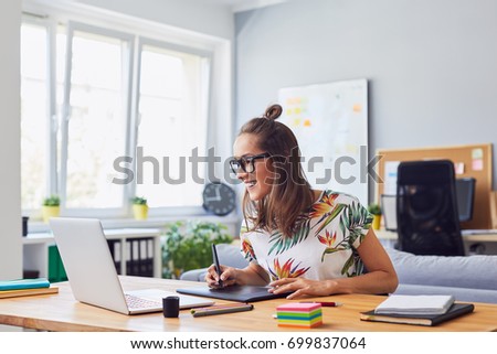 Cheerful attractive young female graphic designer smiling and working at her desk in modern office