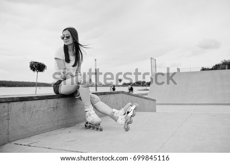 Portrait of an attractive young woman in shorts, t-shirt, sunglasses and roller skates sitting on the concrete bench in the outdoor roller skating rink. Black and white photo.