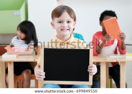 Cute boy holding blank blackboard in classroom, education concept background