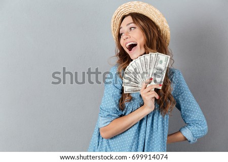 Picture of excited young happy woman standing over grey wall wearing hat holding money. Looking aside.