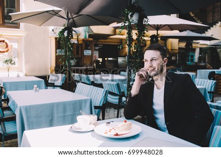 The young businessmen is sitting at the table in restaurant during the short break. He is talking on the phone, smiling and looking straight forward.
