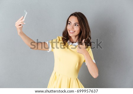 Smiling young girl showing thumbs up gesture while taking a selfie with mobile phone isolated over gray background