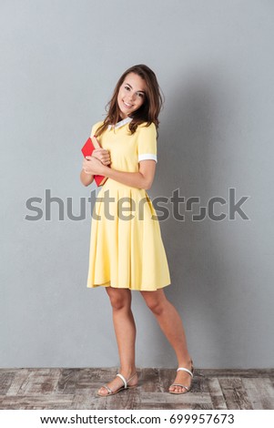 Full length of a casual smiling girl in dress holding book while standing and looking at camera isolated over gray background