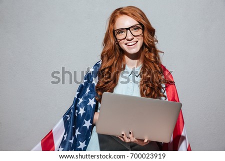 Picture of happy redhead young lady with USA flag using laptop computer. Looking camera.