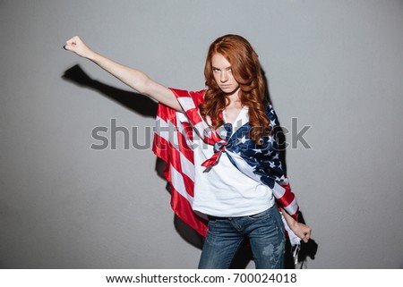 Picture of happy redhead young lady superhero with USA flag. Looking camera.