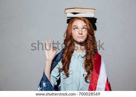 Picture of funny redhead young lady holding book on head wearing USA flag. Looking aside showing okay gesture.