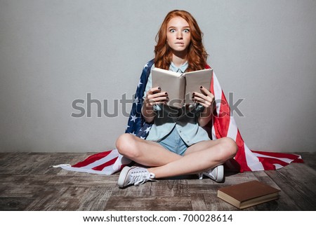 Picture of shocked redhead young lady reading book wearing USA flag. Looking camera.