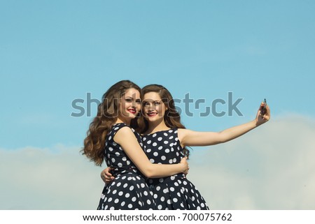 Two beautiful sisters twin girls in identical dresses, with makeup and hairstyle being photographed on the phone against the blue sky