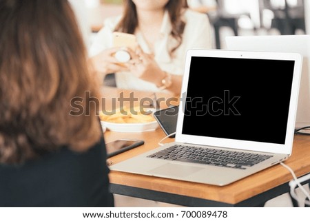 Blank screen laptop place on wooden table. Asian woman working with her friend in coffee shop in selective focus.