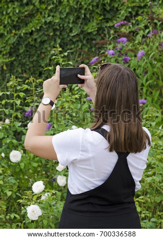 A young girl with a mobile phone photographing a green garden, with a watch on her hands, in a white shirt and a black skirt