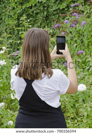 Young girl with mobile phone photographing green garden, with hands and white watch and black skirt