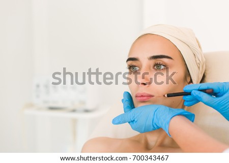 Attractive girl with dark eyebrows, doctor's hands wearing blue gloves drawing punctual lines on the facelines on face.