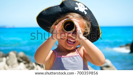 Little happy girl in a pirate hat is playing at the sea looking in a telescope, a background of sea blue water.