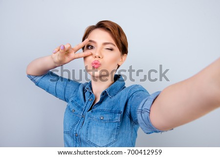 Happy charming young girl taking self portrait showing v-sign and giving a wink, blowing lips