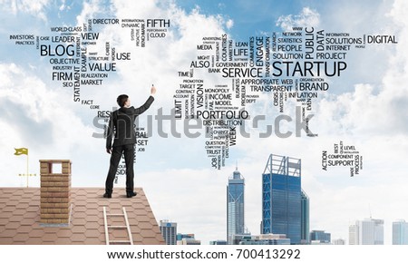 Young businessman standing on house roof and writing business related words. Mixed media