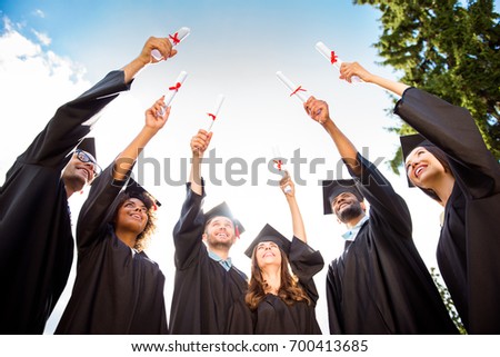Six excited successful graduates in black robes are looking at the certificates in their raised up hands. They did it, passed exams, finished course of studies, got the degree Royalty-Free Stock Photo #700413685