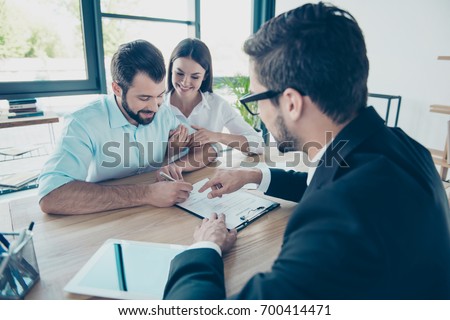 Business meeting. Happy couple is buying new house, young bearded attractive brunet lawyer in a formal wear is presenting contract to sign, they are in his office