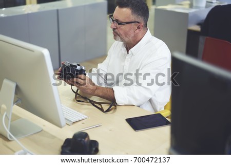 Professional male employee in eyeglasses sitting at computer and making photos for productive work.Mature man in optical spectacles taking pictures on stylish vintage camera in coworking space