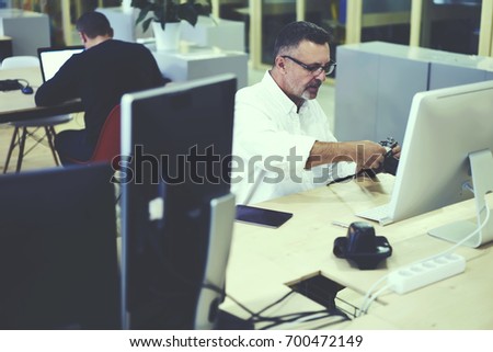 Professional photographer 40 years old in optical spectacles holding vintage camera in hands and making settings for editing during working process in office interior sitting at desktop with computer