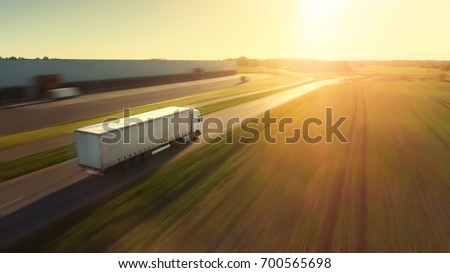 Aerial Follow Shot of White Semi Truck with Cargo Trailer Attached Moving Through Industrial Warehouse, Rural Area. Sun Shines and the Sky Are Blue. Blur motion.