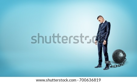 A depressed businessman on blue background stands with a lowered head while chained to an iron ball. Business world restrictions. Credit burden. Financial obligations.