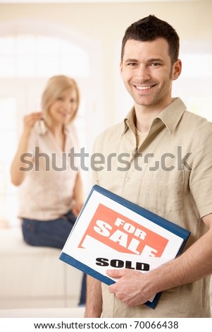 Happy man standing with for sale sign, girlfriend in background holding keys of house.?