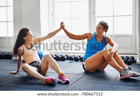 Two smiling young female friends in sportswear sitting together on the floor of a gym high fiving each other Royalty-Free Stock Photo #700667512