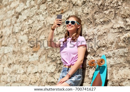 lifestyle, summer, technology and people concept - smiling young woman or teenage girl in sunglasses with longboard and smartphone taking selfie over stone wall outdoors