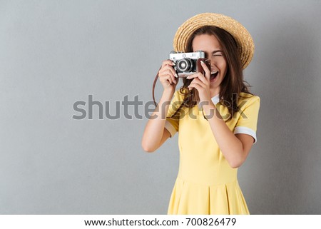 Young pretty girl in hat taking picture with vintage camera while standing isolated over gray background