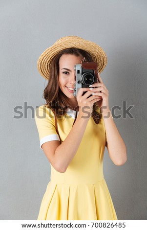 Smiling pretty girl in hat taking picture with vintage camera while standing isolated over gray background
